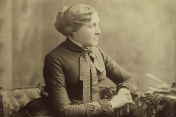 A photo of Louisa May Alcott in profile with her arms up on the arm rest of a chair, hands folded. She is only visible from the waist up.